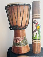 Photo showing the African wooden drum with rope-tensioned skin and detailed beadwork, alongside the cylindrical rain stick with colorful parrot painting.
