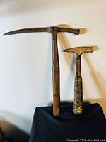 Two antique cast iron hammers side by side on black cloth. Larger hammer features long pick and wooden handle. Smaller hammer features leather wrapped handle.