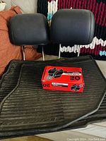 Three black rubber floor mats stacked with two black leather tilting headrests and a red box of brake parts on top