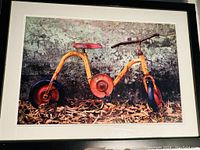 Full view of the framed and matted photograph of a vintage yellow tricycle with rust and dry leaves in front of a wall.