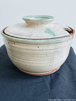 Side view of handmade pottery bowl with lid, cream with green accents and a visible crack on the rim edge.