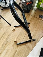 Black tubular steel rear motorcycle stand with two hooks for rear axle, photographed on a wood-like floor.