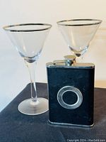 Two silver-rimmed martini glasses and a black leather-covered flask with circular metal plate on front, standing side by side against a light background.