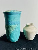 Front view of two pottery vases on black surface, turquoise vase on left with lid resting next to it, beige smaller vase on right.