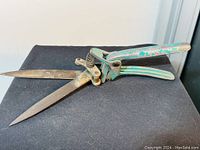 Side view of vintage metal pruning shears featuring long blades and green paint on handles, showing wear and rust.
