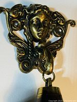 Close-up photo of the brass ornate Victorian hanging bell showing detailed woman's face and decorative wings above the bell.
