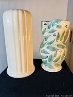 Photo showing cream ceramic vase with vertical ridges and ceramic jug decorated with green leaves and blue berries on black fabric background