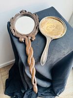 Two vintage hand held mirrors placed side by side on a dark cloth, one wooden with carved ornate frame and twisted handle with mirror, the other sterling silver with empty frame.