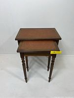 Pair of rectangular vintage nesting tables, stacked with smaller table under larger. Both tables have brown leather tops with decorative gold borders and wooden turned legs.