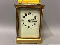 Front view of the carriage clock showing the white dial with black numerals and two black hands, brass casing with some patina and wear.