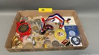 Wide view of assorted commemorative pins and medals in cardboard box showing various metals, ribbons, and emblem styles.