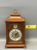 Full front view of the wooden Franz Hermle mantle clock, showing brass handle on top and feet, clear glass front with ornate metal face and Roman numerals.