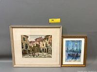 Frontal view of both watercolor paintings framed and leaning against a gray wall showing the full lot