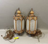 Pair of hexagonal vintage hanging light fixtures with gold-toned ornate metal frames and frosted glass panels seen front view with coiled chains and cords placed in front.