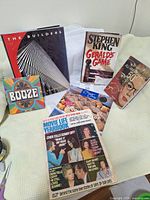 Photo showing all 5 hardcover books and the vintage Movie Life Yearbook magazine arranged on a bed. Visible titles include The Builders, Gerald's Game, All About Aging, and BOOZE. The magazine front cover highlights celebrity gossip and soap opera stories from the 1950s.