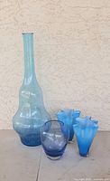 Tall slender vase with light blue tint and silver flecks, three small matching blue vases with ruffled edges, and medium rounded blue vase arranged on a surface against textured wall.