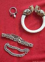 Four costume jewelry bracelets on red background including peacock beaded bracelet, silver-tone geometric link bracelet, chunky chain bracelet, and open adjustable bangle with charms.