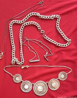 Photo of five costume jewelry necklaces on red background, showing large thick chain, medallion necklace, and delicate chains with letter pendants.