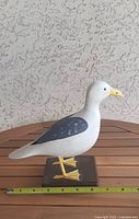 Side view of carved wooden seagull showing white body, dark blue wings, yellow beak and feet, standing on square base.