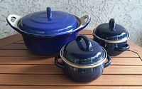 Three blue enamel cast iron casserole dishes on wooden surface against textured wall. Large casserole has white interior shown with lid off. Two smaller casseroles have dark blue enamel inside and out with matching lids.