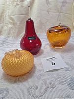 Photo showing the three glass fruit paperweights: a red pear with clear stem labeled 'KAKABAT', an amber apple, and a textured gold apple.
