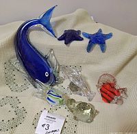 Photo of all seven glass sea life figurines arranged on cream textured fabric, showing large blue fish, starfish, angel fish, turtle, and dolphin.