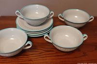 Eight white porcelain broth bowls with two handles each, placed on wooden table along with eight matching saucers, all with celadon green rims and soft textured patterns.