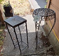 Two exterior metal plant stands on a concrete patio, square stand with leaf design and round stand with swirl pattern.