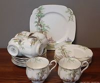 Photo showing stacked dinner plates, salad plates, teacups, and saucers with Royal Albert Greenwood Tree pattern featuring trees, cottages and gold trim.