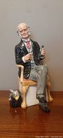 Front view of the Royal Doulton The Doctor figurine; seated elderly male doctor holding green bottle with medical bag nearby.