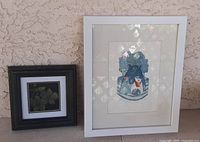 Two framed artworks shown side by side against an outdoor textured wall. Left is smaller framed leaf art by Gail Reed, right is larger Edie Miller 'Fat Cat' numbered print in white frame.