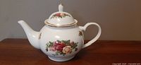 Side view of ornate white porcelain teapot with floral rose pattern and gold trim showing rounded body, spout, and handle.