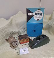 Philishave and Birks boxes, cast tape dispenser, pencil sharpener, and Twinplex razor sharpener displayed on light fabric
