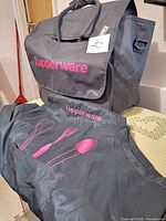 Photo of black fabric Tupperware tote bag with handles and black apron with pink graphics displaying the Tupperware brand name and kitchen utensils.