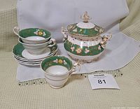 Photo showing the full set including sugar bowl with lid, three cups and saucers, and three small bowls arranged on a beige textured surface with a white cloth background.