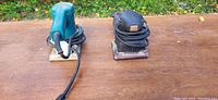 Two electric sanders placed on a wooden surface, showing the top and side views of both units with cords wrapped.