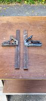 Photo shows two vintage metal sliding squares with attached wooden rulers on a wooden surface, demonstrating condition and details.