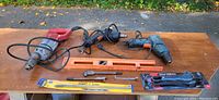 Overview of three electric drills, a broken orange level, a packaged saw blade, and drill accessories on a wood table.
