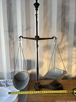 Full view of antique metal balance scale with two metal pans hanging from chains, placed on wooden surface with measuring tape for scale.