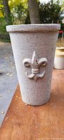 Outdoor photo of tall, tapered cast iron vase with textured speckled white finish and raised fleur-de-lis decoration on the front.
