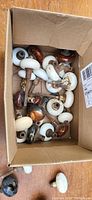 Box filled with various white, brown, and black porcelain door knobs, some attached to metal spindles.