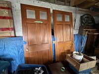 Pair of two solid wood French doors leaning against wall, showing full front with four glass panes each and lower wood panels, blue tarp behind.