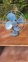 Front view of the vintage blue desk fan on a wood surface with foliage background.