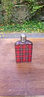 Front view of vintage red plaid fabric covered flask with silver-tone cap on wooden surface with outdoor background