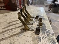 Four brass hand rail brackets positioned upright and four brass clamps laid out on a work surface with scattered tools and materials in the background.