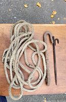 Photo shows a coiled natural fiber rope and two rusty metal hooks placed on a wooden surface outdoors.