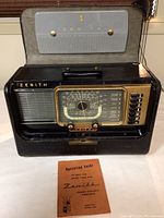 Front view of the Zenith Trans-Oceanic radio inside the carrying case with the open lid, showing the large tuning dial, buttons, carrying handle, and the operating guide placed in front.