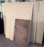 Stack of Masonite panels of varying sizes leaning against wall, including a wood-grain finish panel.