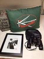 The green Mercury Kiekhaefer pillow, black Tasco binoculars with carrying case, aluminum fuse box, and part of the black and white framed photo arranged on a surface.