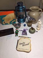 Photo showing entire lot on table including painting, lantern, ceramic pitcher, bird figurines, plastic Madonna, elf figurine, bag of sea glass, old metal bicycle bell, tiny rifle souvenir, and vintage cookbook.